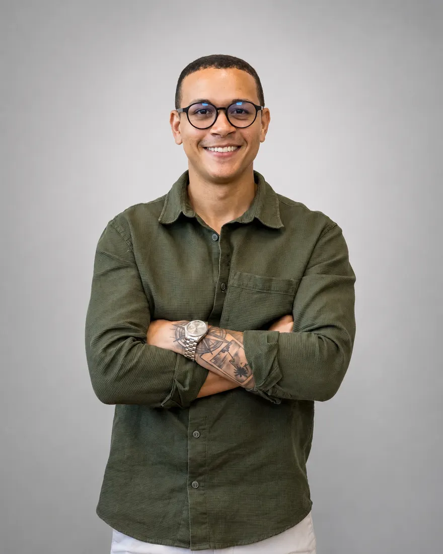 Brotherson Américo, de óculos e camisa verde, sorri de braços cruzados.